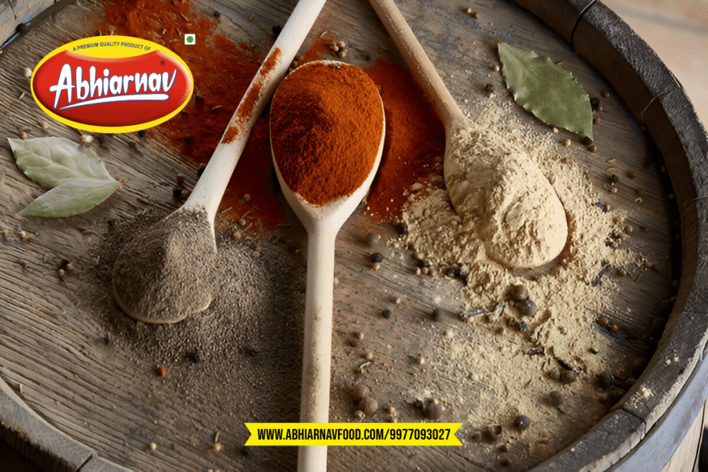 Abhiarnav Food Products seasoning spices display featuring three wooden spoons with red orange gray and beige spice powders on rustic wooden background with bay leaves